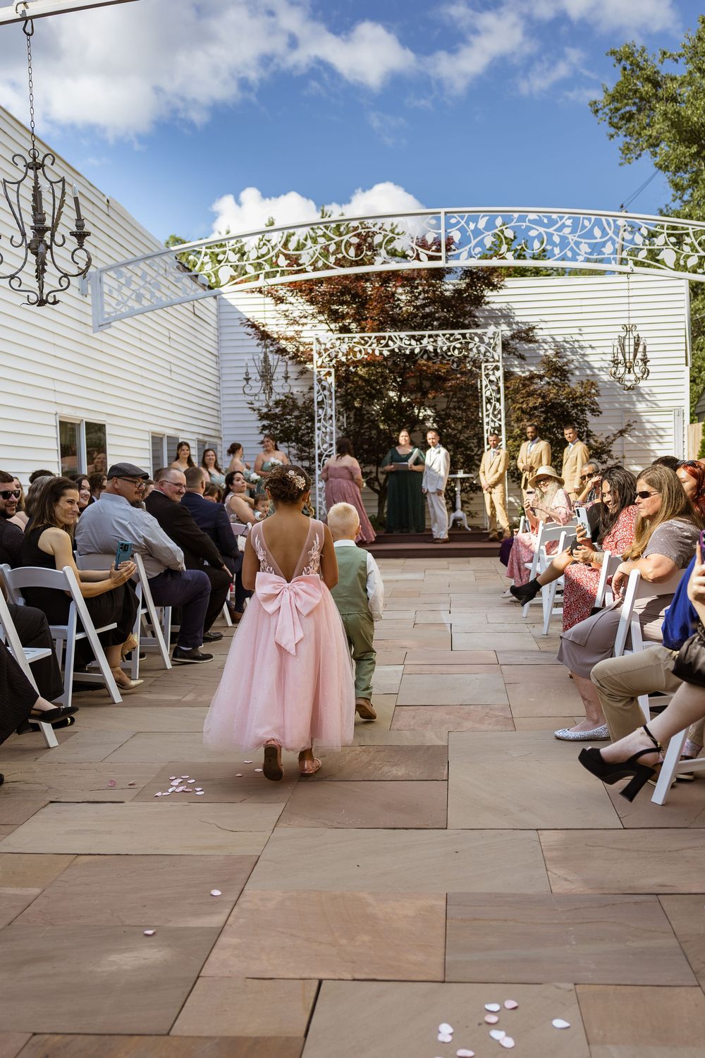 Flower girl in pink dress walks down outdoor wedding aisle toward floral arch altar with guests seated on both sides.