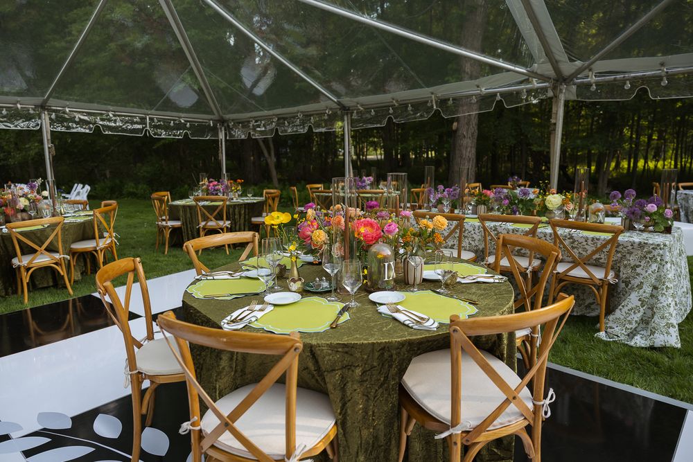 Elegant outdoor wedding reception under clear tent with rustic wood chairs, round tables, green linens, and colorful floral centerpieces.