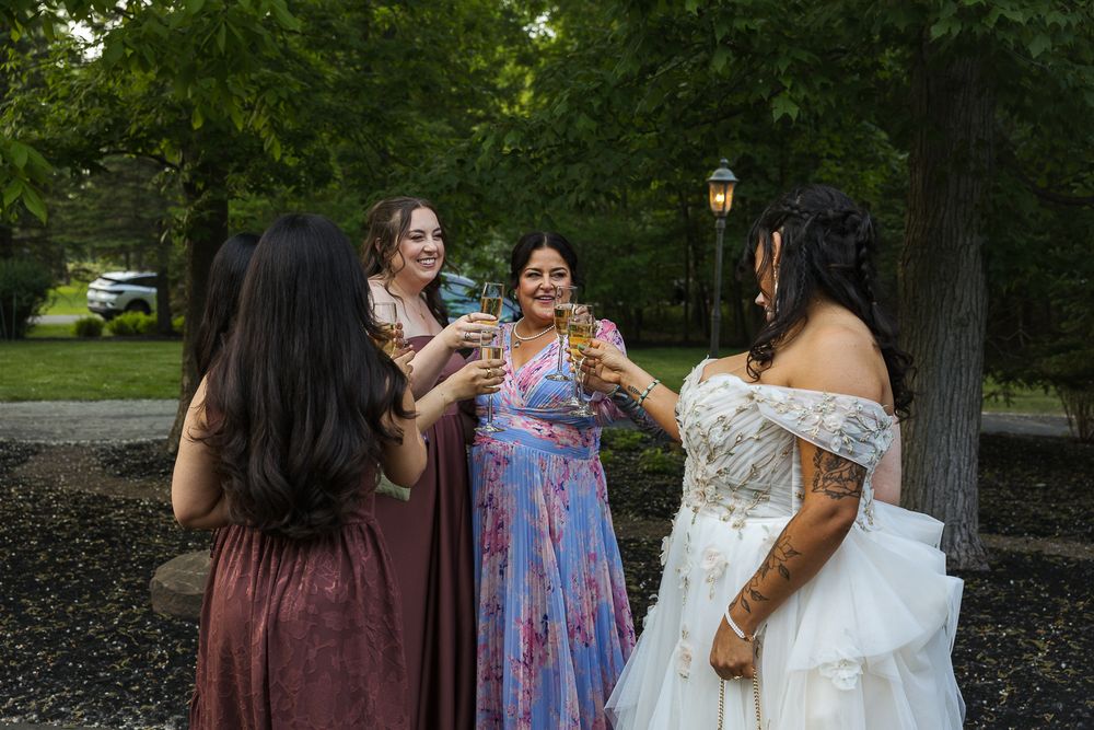 Bride in white gown toasting champagne with bridesmaids in colorful dresses outdoors among lush green trees.