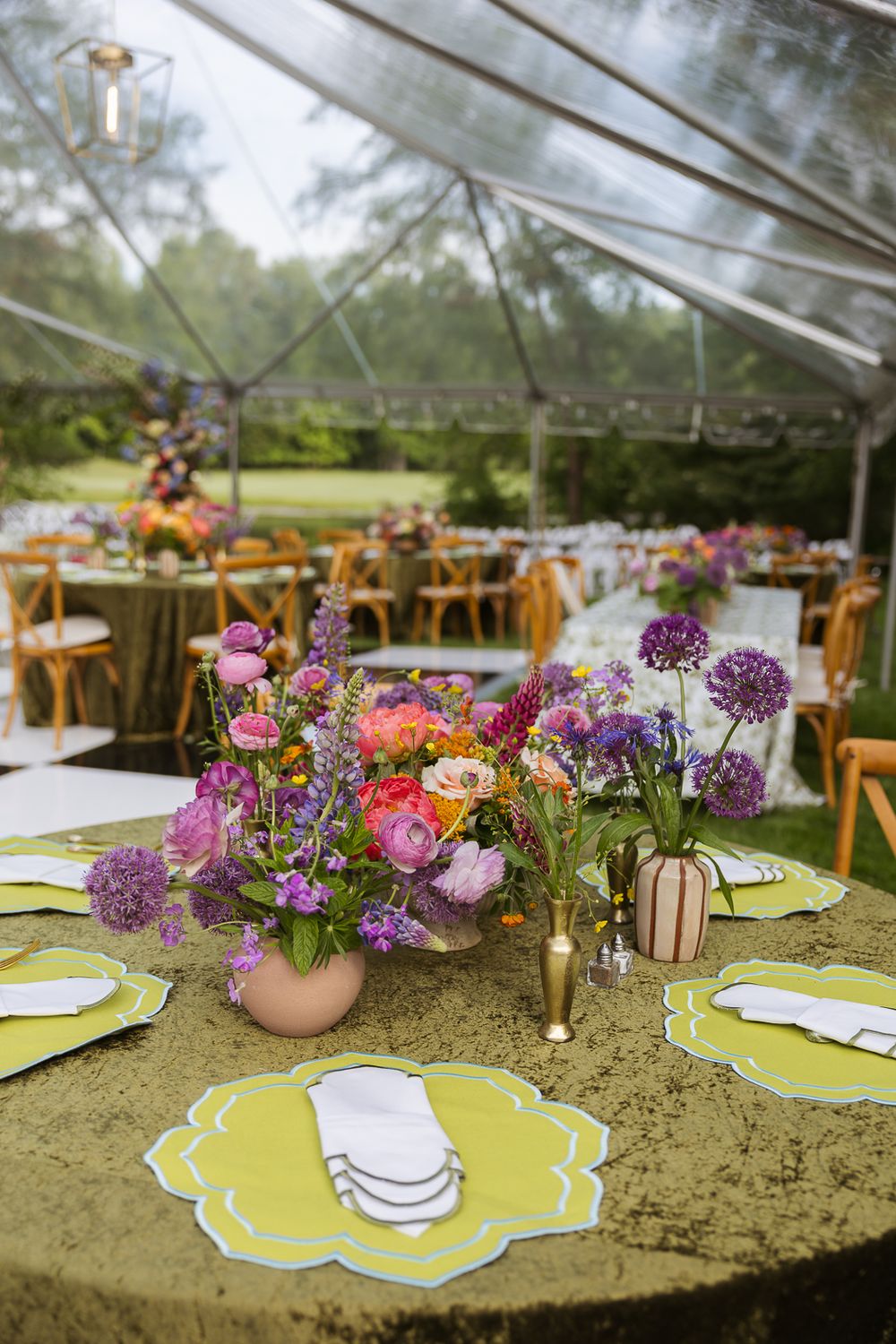 Colorful outdoor wedding reception under clear tent with vibrant floral centerpieces, green tablecloths, and yellow placemats.