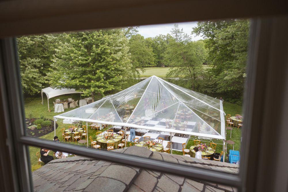 Aerial view through window of elegant outdoor wedding reception under clear tent with round tables on lush green lawn.