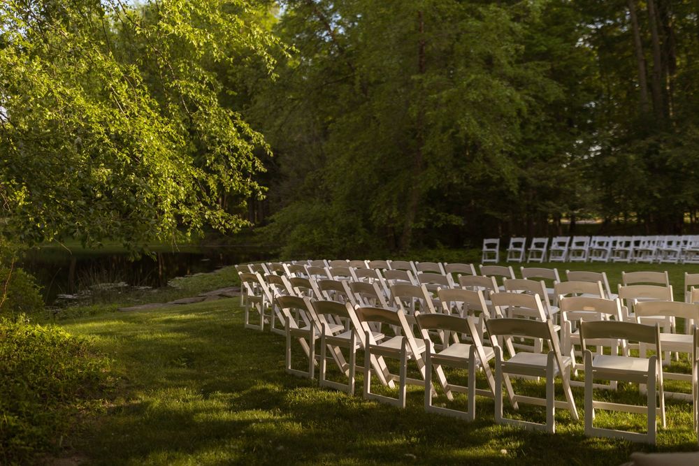 Rows of white folding chairs arranged outdoors on green grass surrounded by lush trees for an elegant garden ceremony.