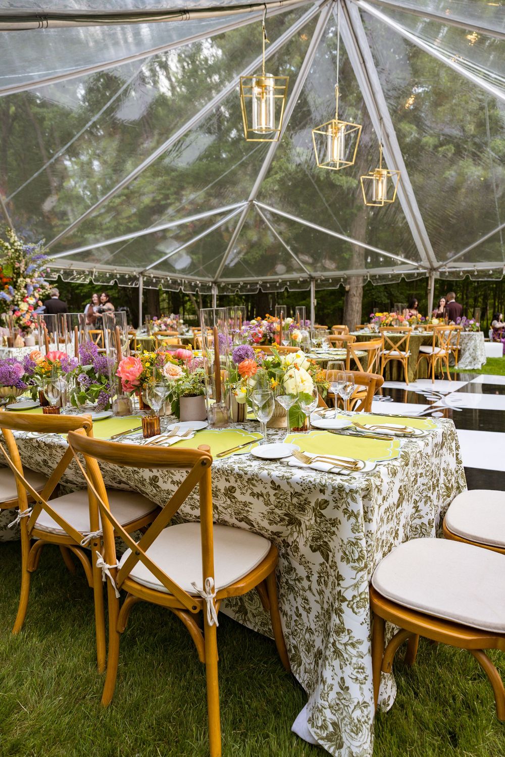 Elegant outdoor wedding reception under clear tent with floral centerpieces, gold lanterns, and rustic wooden cross-back chairs.
