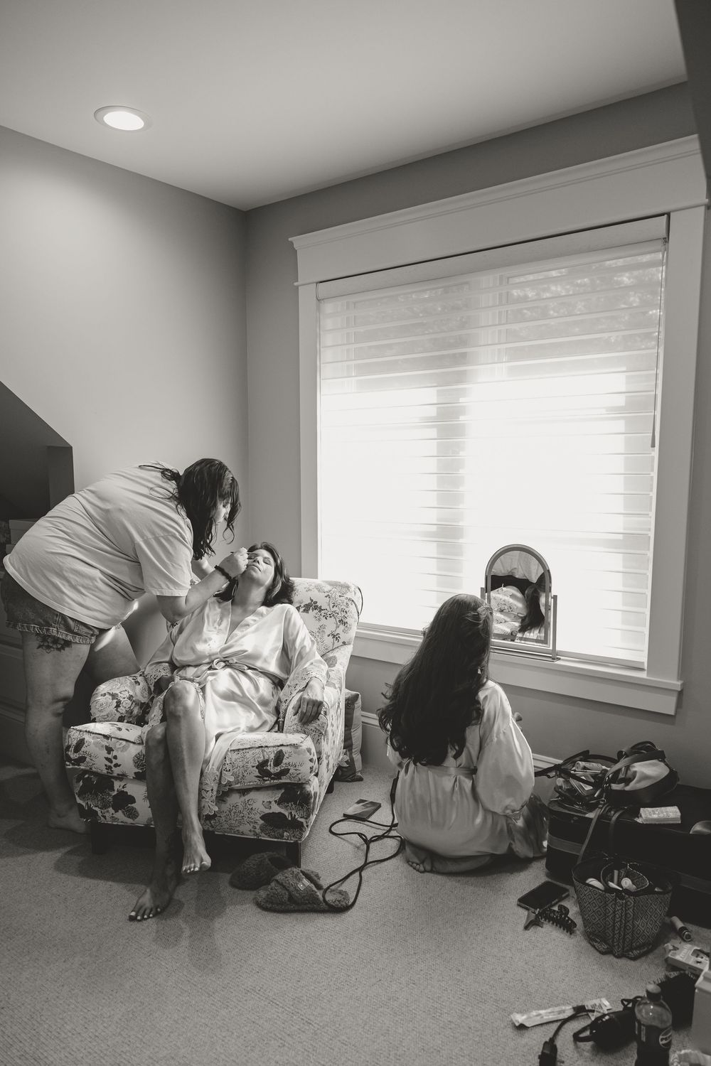 Black and white photo of bride getting hair and makeup done by stylists in a bridal suite before wedding.