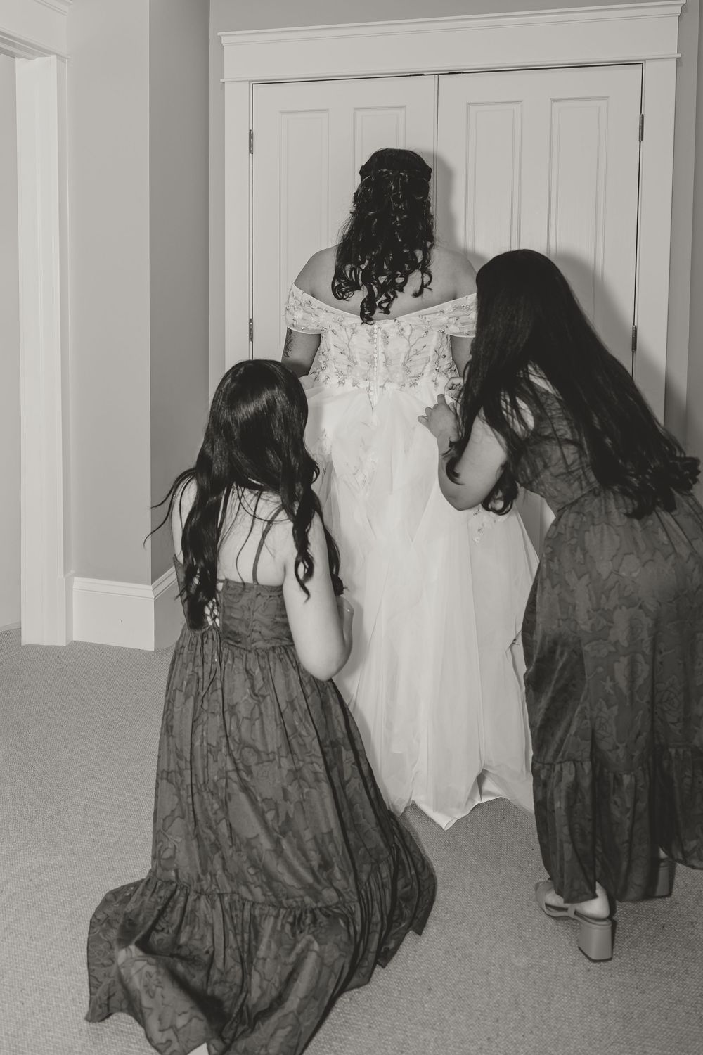 Black and white photo of a bride in a lace gown being assisted by two bridesmaids in long dresses.