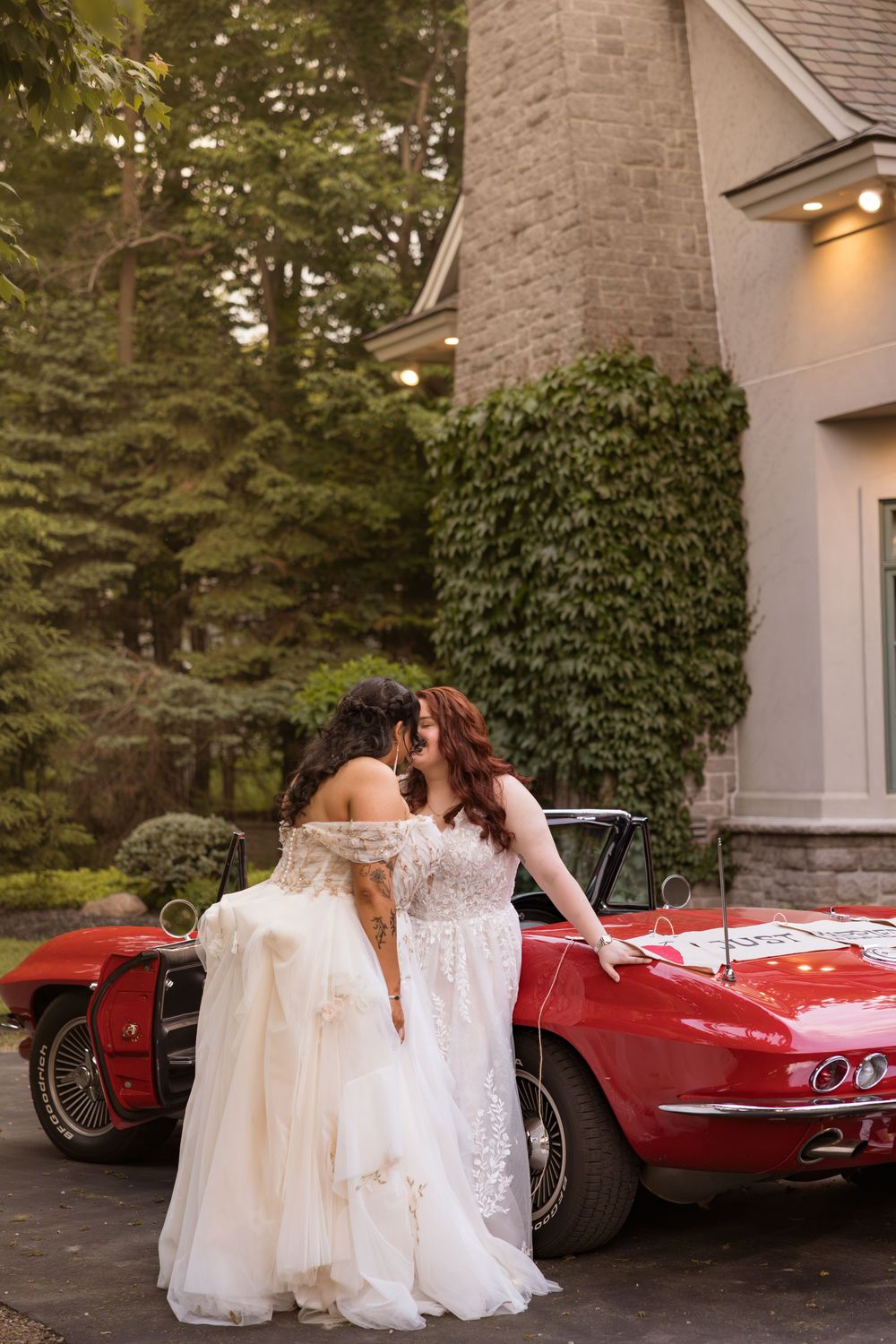Two brides in white gowns share a romantic moment leaning against a classic red Corvette convertible.