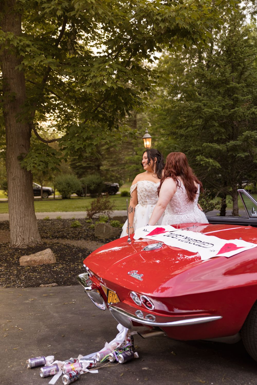 Two brides in white gowns lean against a classic red Corvette convertible decorated with wedding signs.