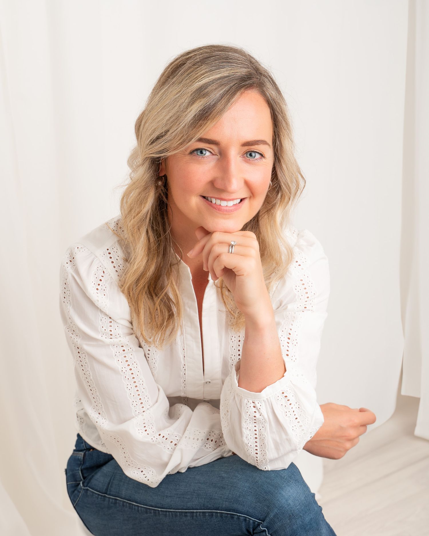 Smiling blonde woman in white eyelet blouse and jeans poses confidently in bright studio setting.