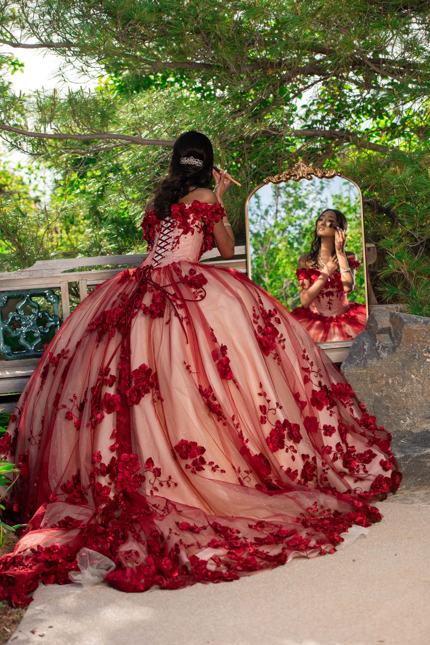 Girl in stunning red floral ball gown viewing reflection in ornate mirror in lush garden.