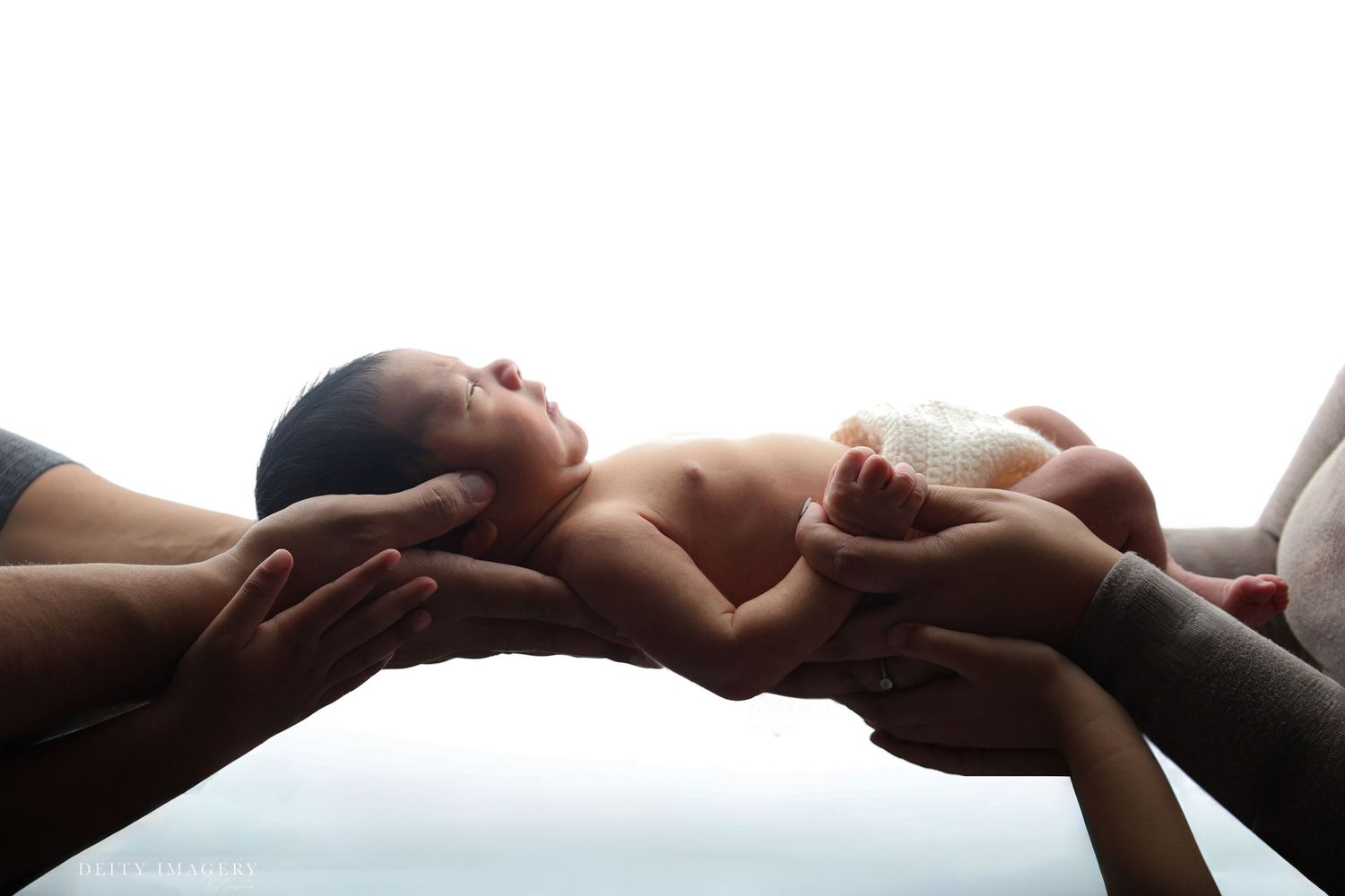 Newborn baby cradled by multiple hands against a bright white background.