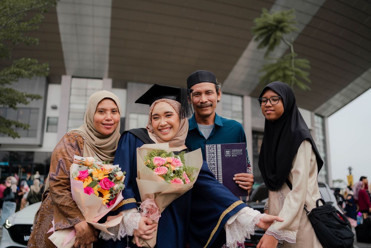 Muslim family celebrating graduation, graduate in blue cap and gown holds flowers and diploma with proud relatives.