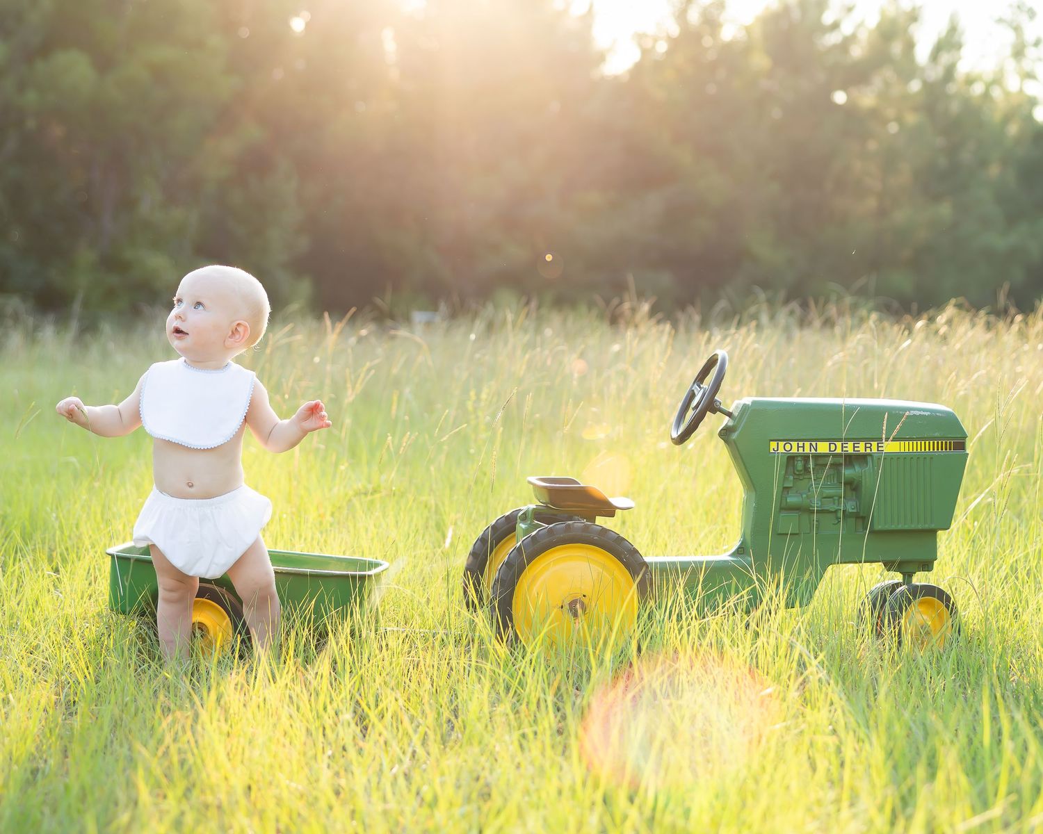 Toddler in denim overalls standing on a green John Deere toy tractor in a grassy field at golden hour.