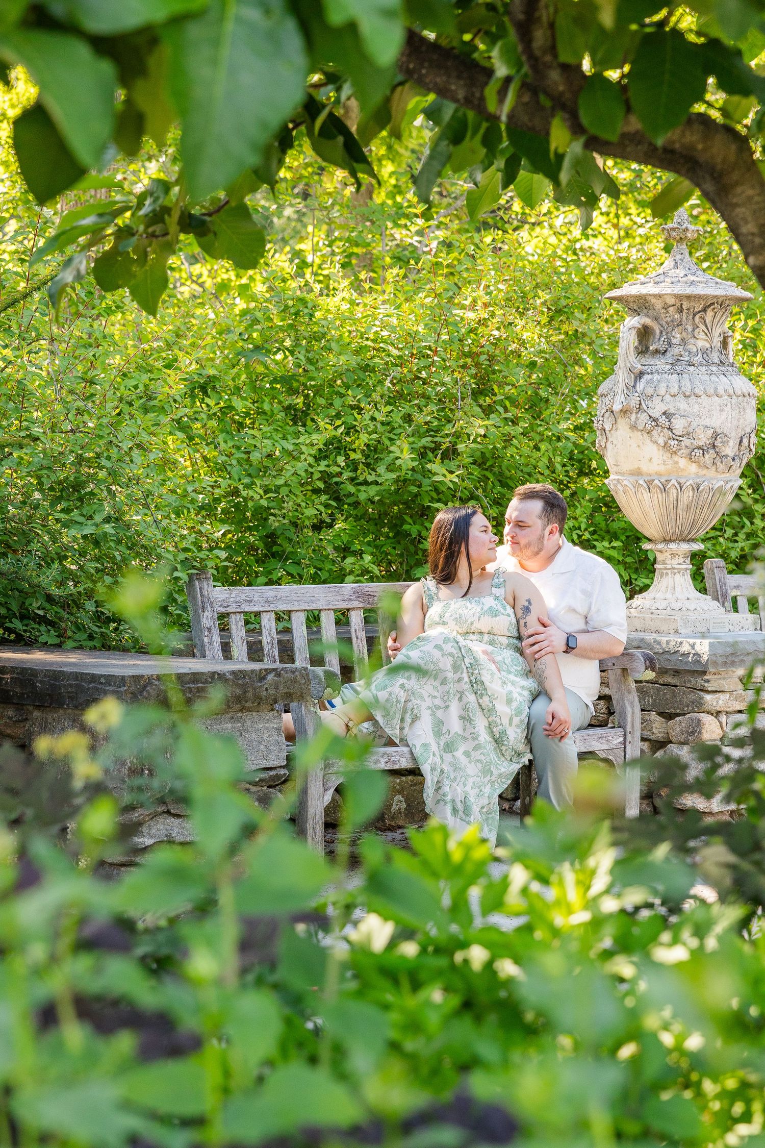 Couple sitting on a wooden bench in a lush garden, smiling at each other near a stone urn.
