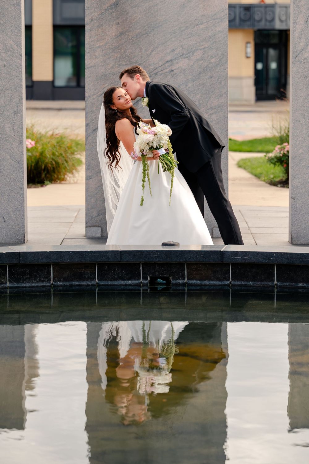 Bride and groom sharing a kiss near a reflective pool, with the couple's reflection visible in the water below.