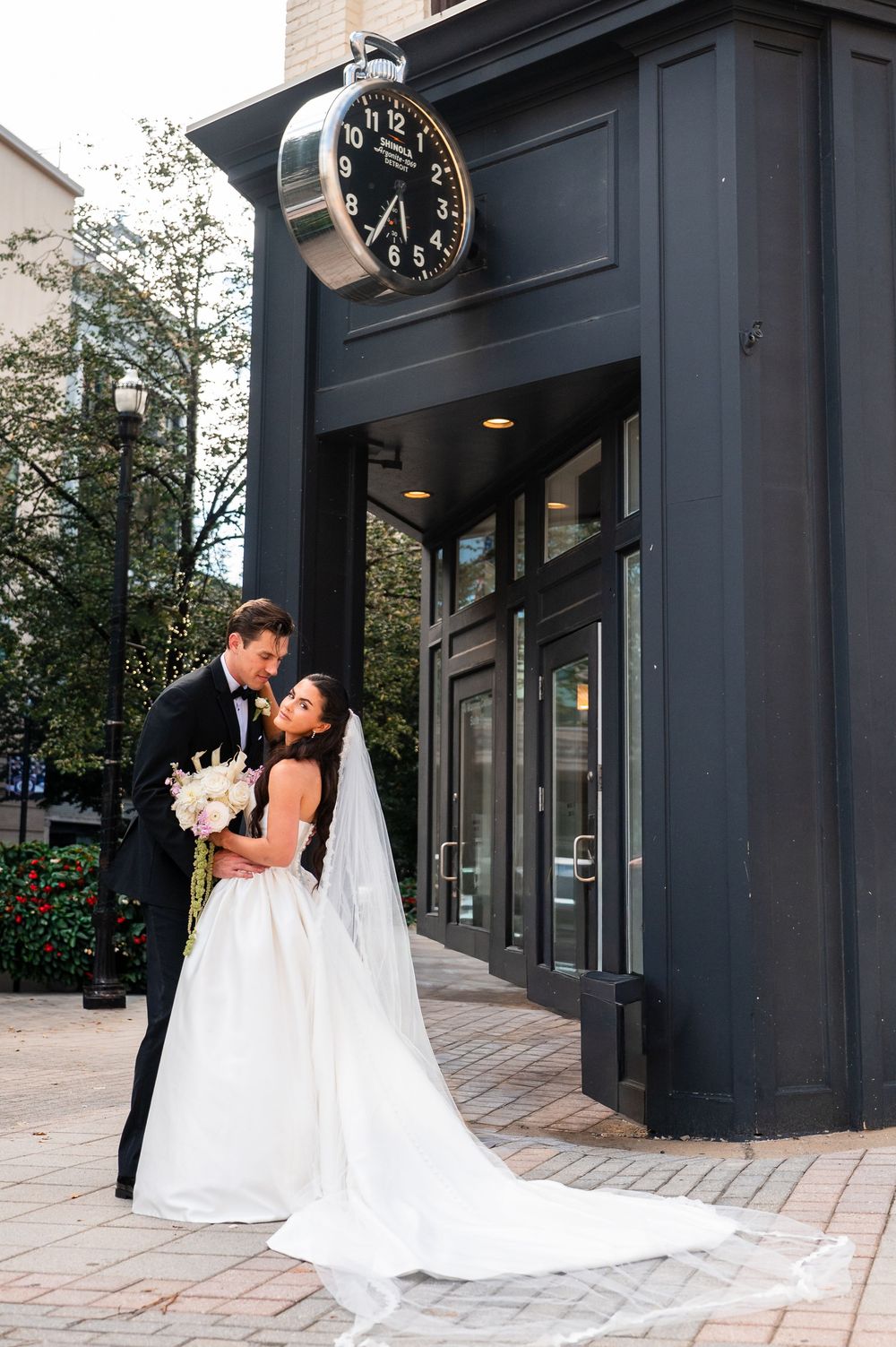 Bride and groom share a romantic moment outside a building with a large clock, bride holding a white bouquet.