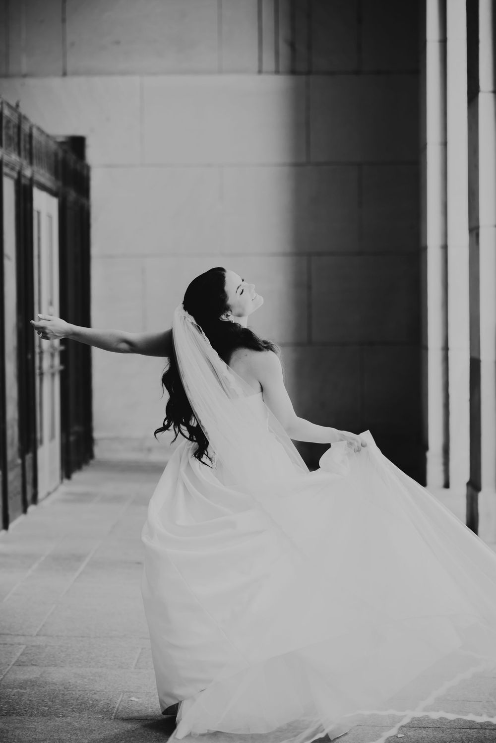 Black and white photo of a bride in a flowing gown and veil, arms outstretched, posing gracefully outdoors.