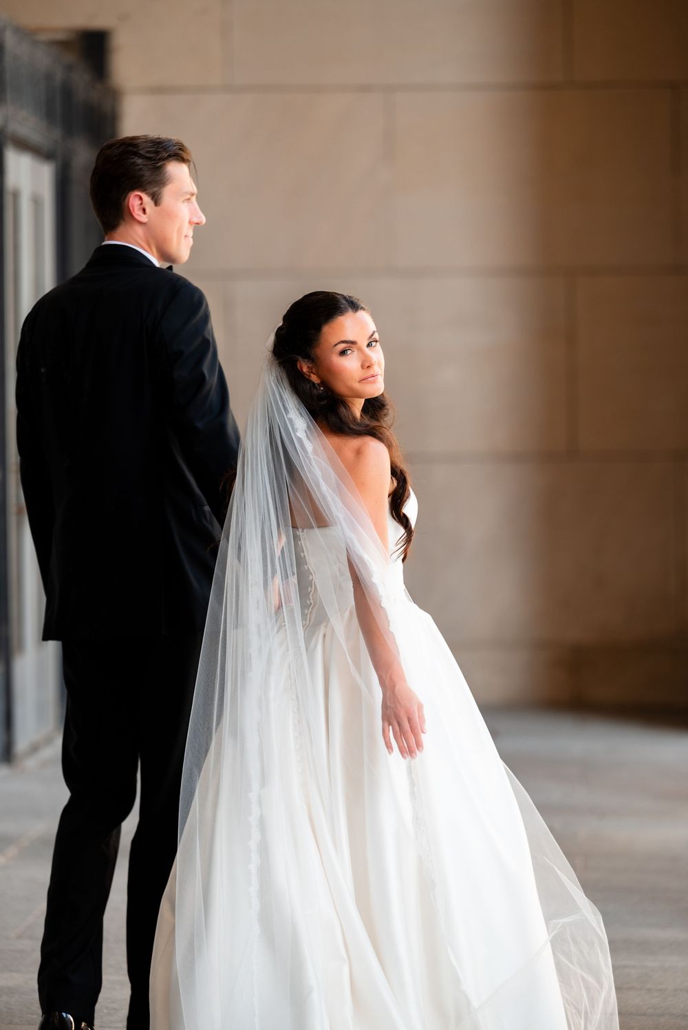 Bride in white ballgown with veil stands beside groom in black tuxedo during elegant outdoor wedding portrait.