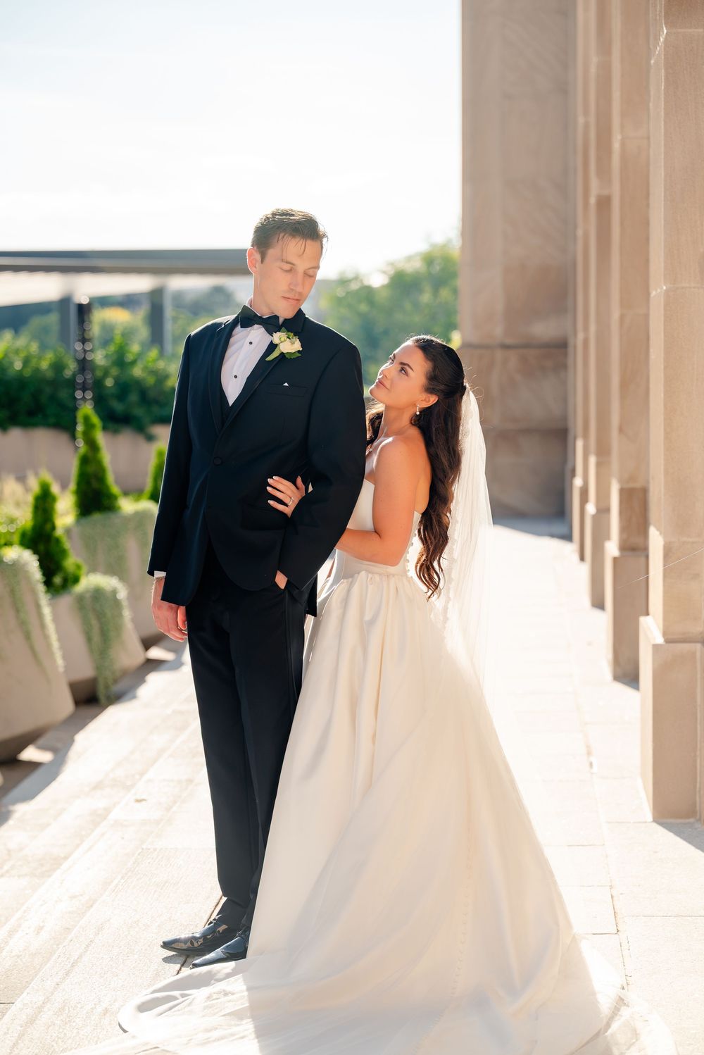 Bride in white gown and groom in black tuxedo share a romantic moment outdoors by stone columns on their wedding day.