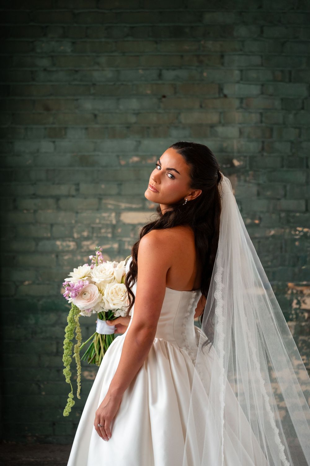Bride in strapless white gown and veil holds floral bouquet, posing elegantly against a green brick wall.