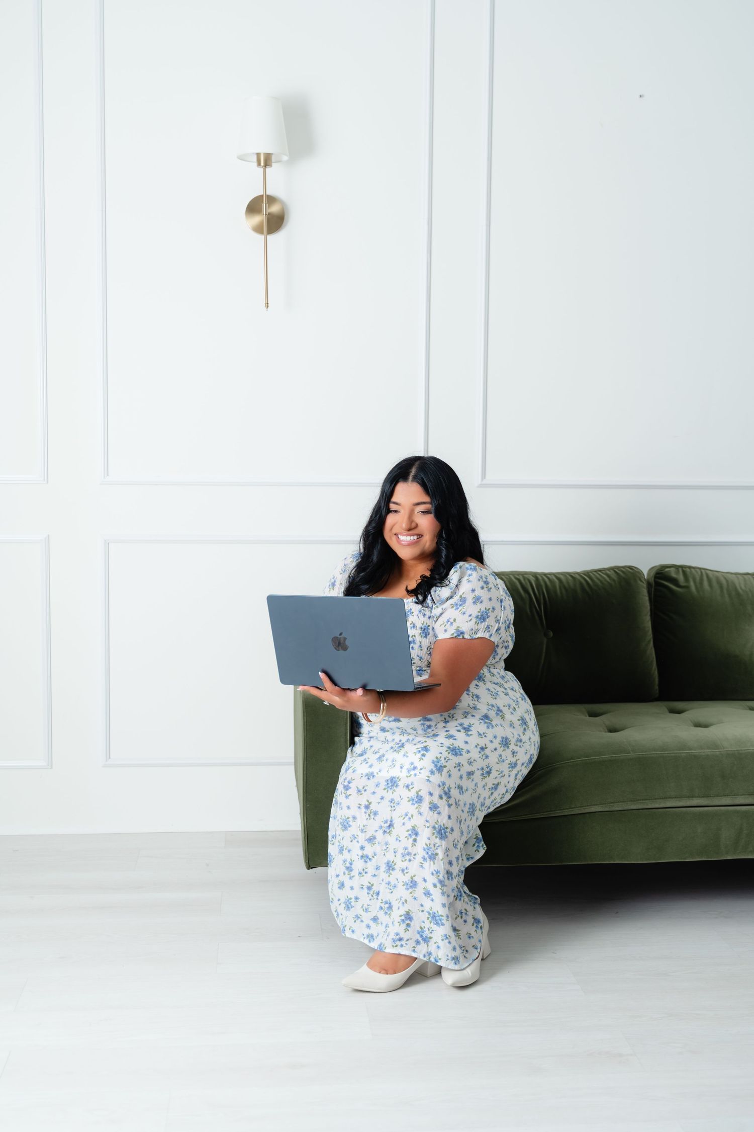 Smiling woman in floral dress sitting on green velvet sofa holding a MacBook laptop.
