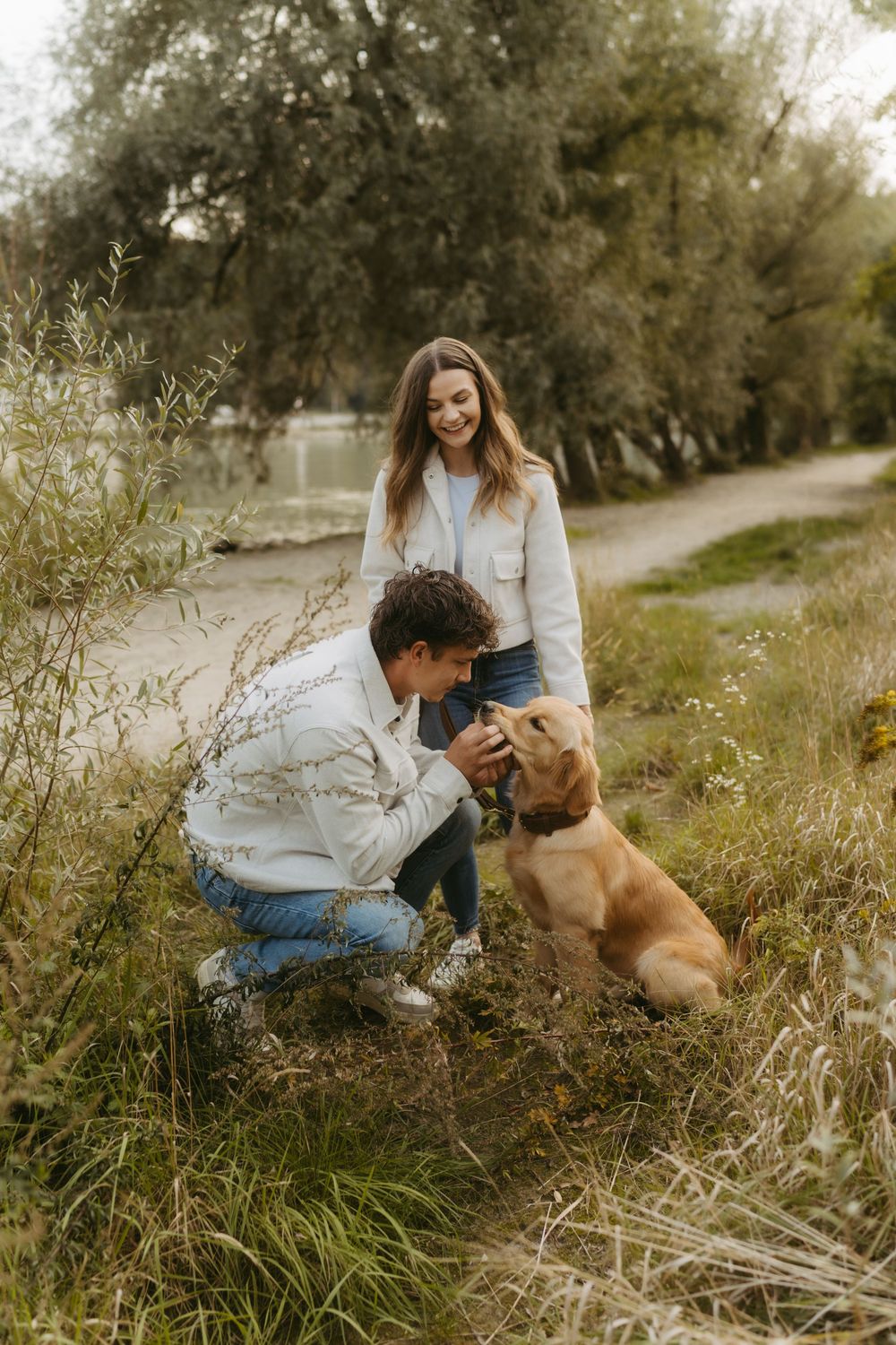A smiling couple interacts with a golden retriever puppy outdoors on a nature path surrounded by greenery.