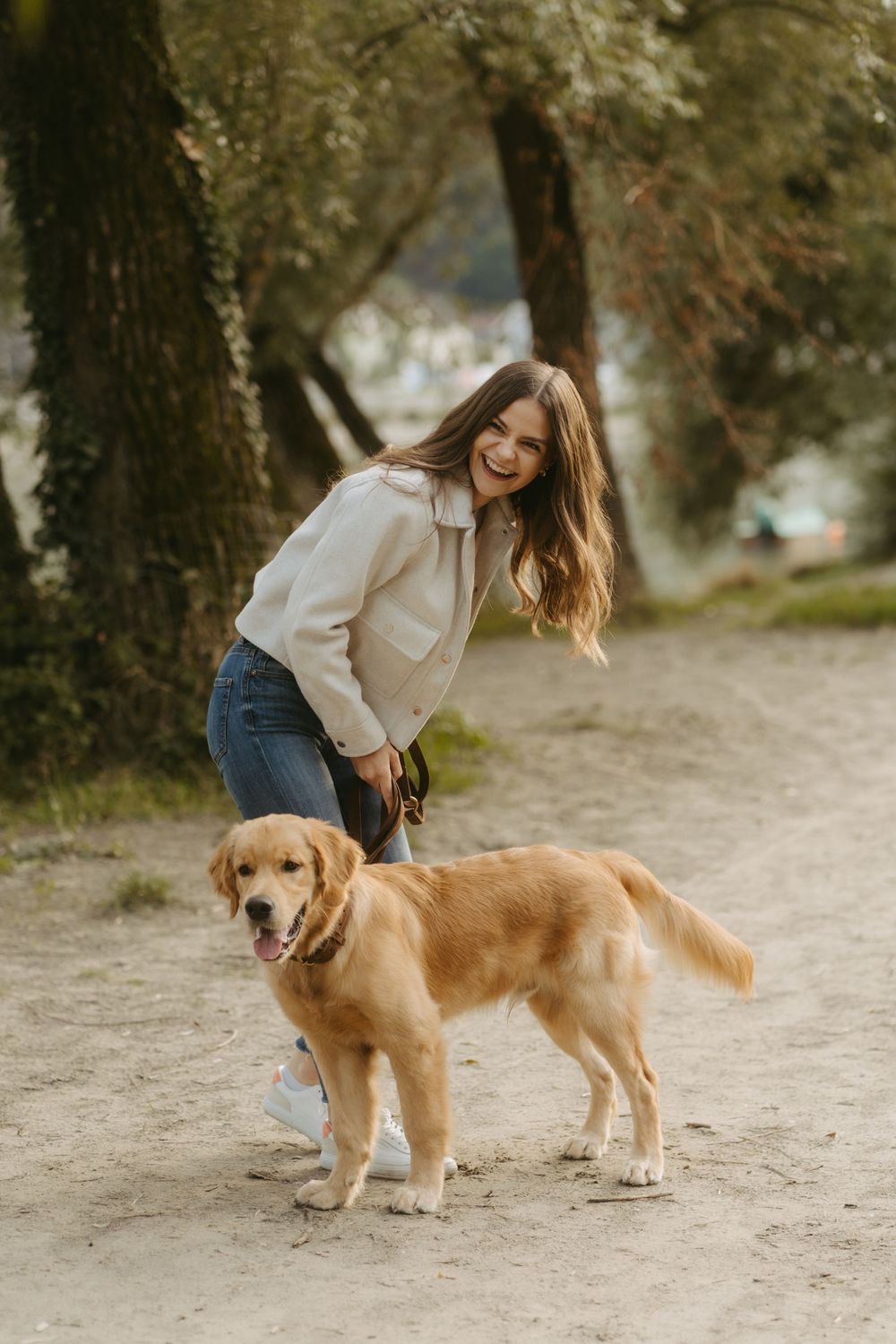 Young woman in white jacket smiling with golden retriever dog on sandy path in forest park.