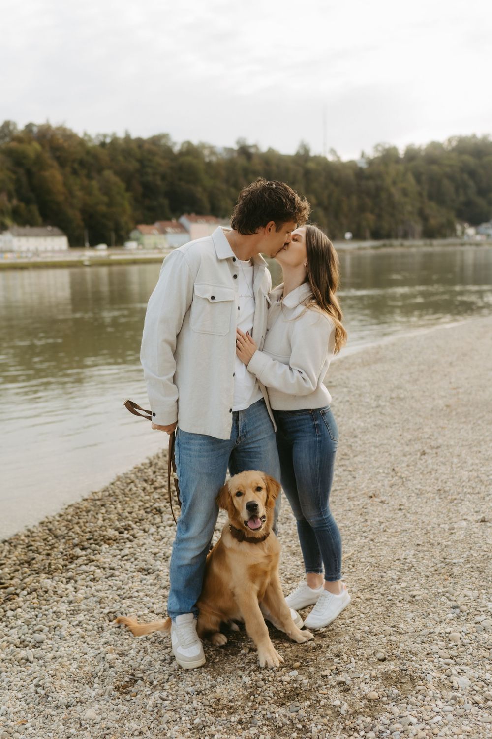 Couple kissing by a lakeside shore with their golden retriever dog sitting in front, trees in background.