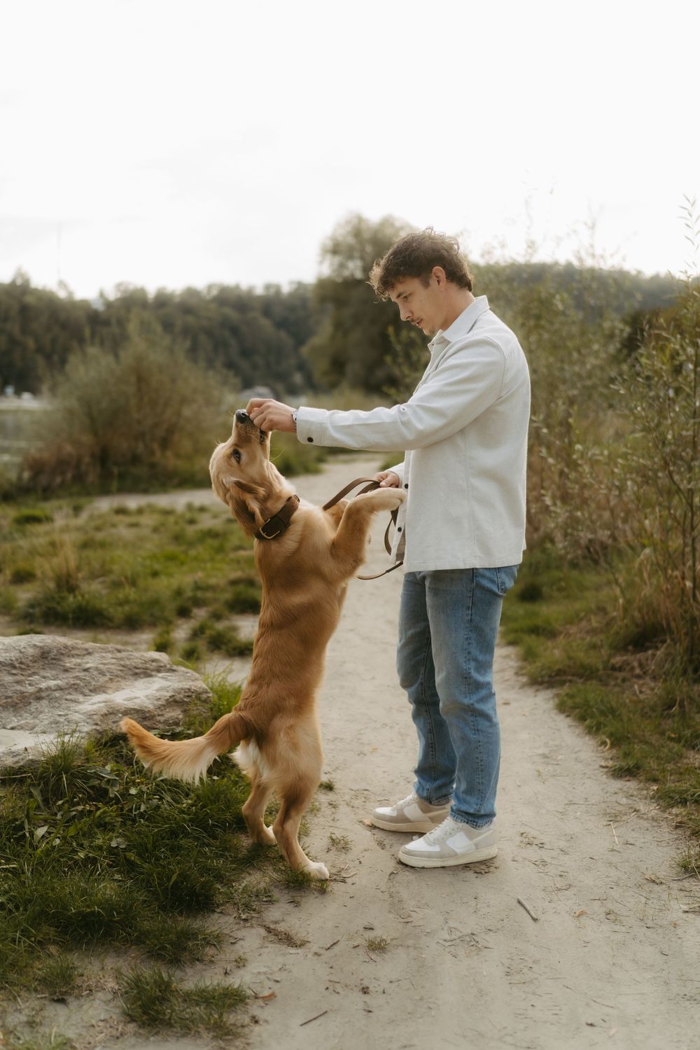 A young man in a white jacket and jeans plays with a golden retriever on a sandy path in a park.