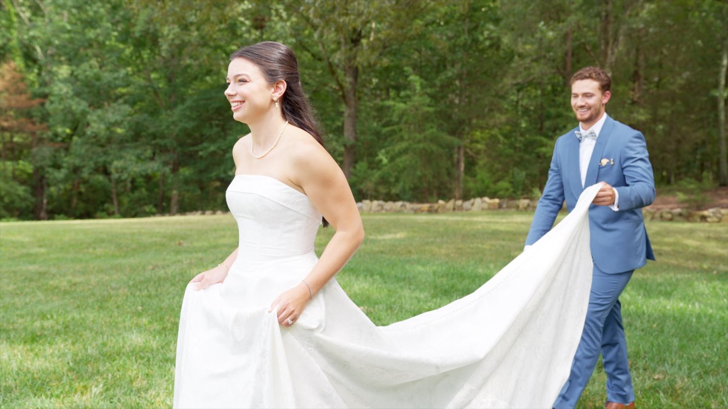 Bride in white strapless gown walks outdoors as groom in blue suit holds her train on lush green lawn.