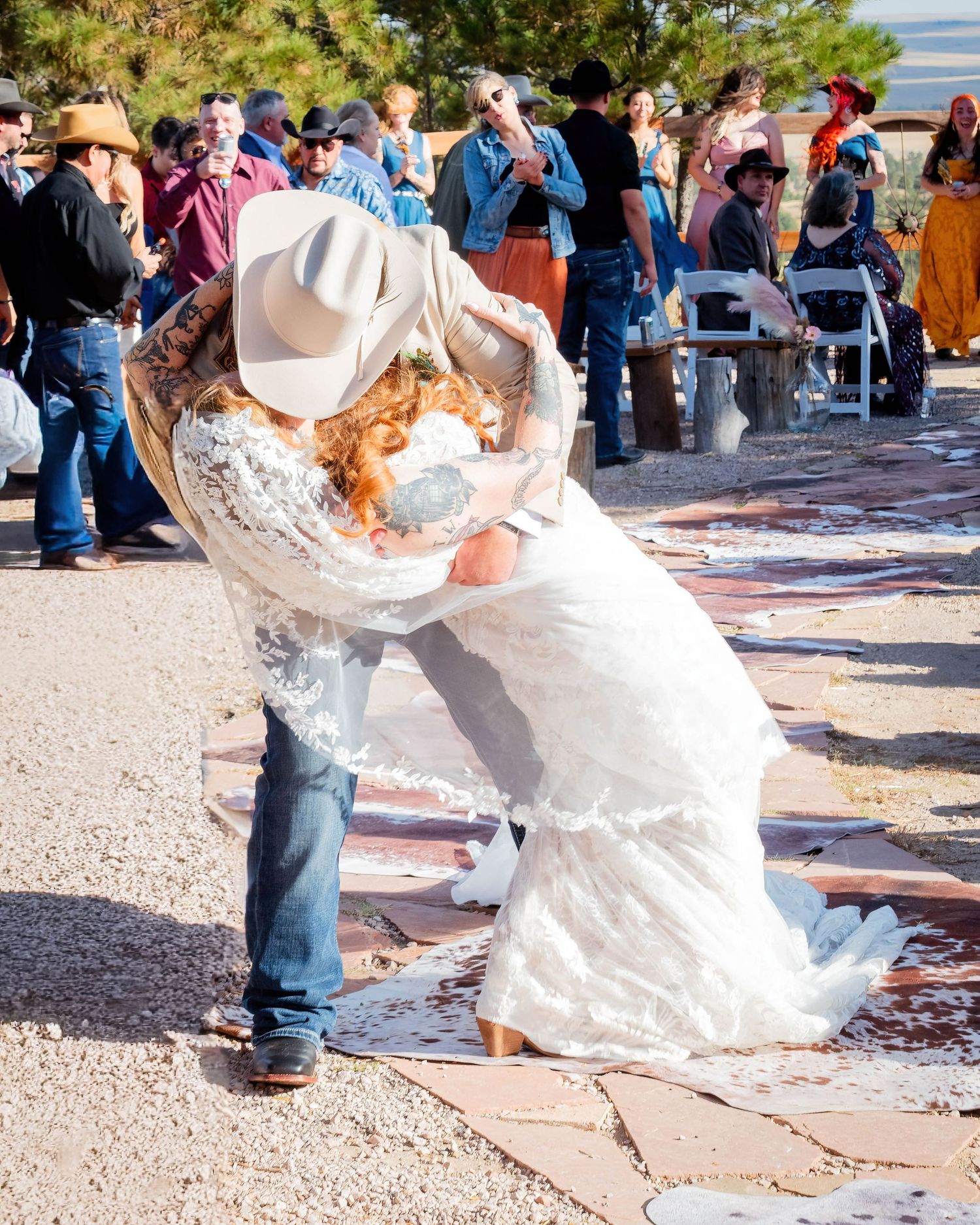 Couple sharing a romantic dip kiss at outdoor western wedding, bride in lace gown, groom in cowboy hat and jeans.