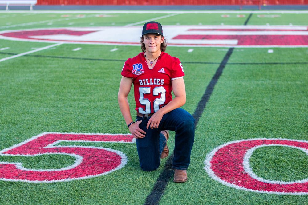 Student athlete in Billies #52 jersey kneeling on football field at the 50-yard line.