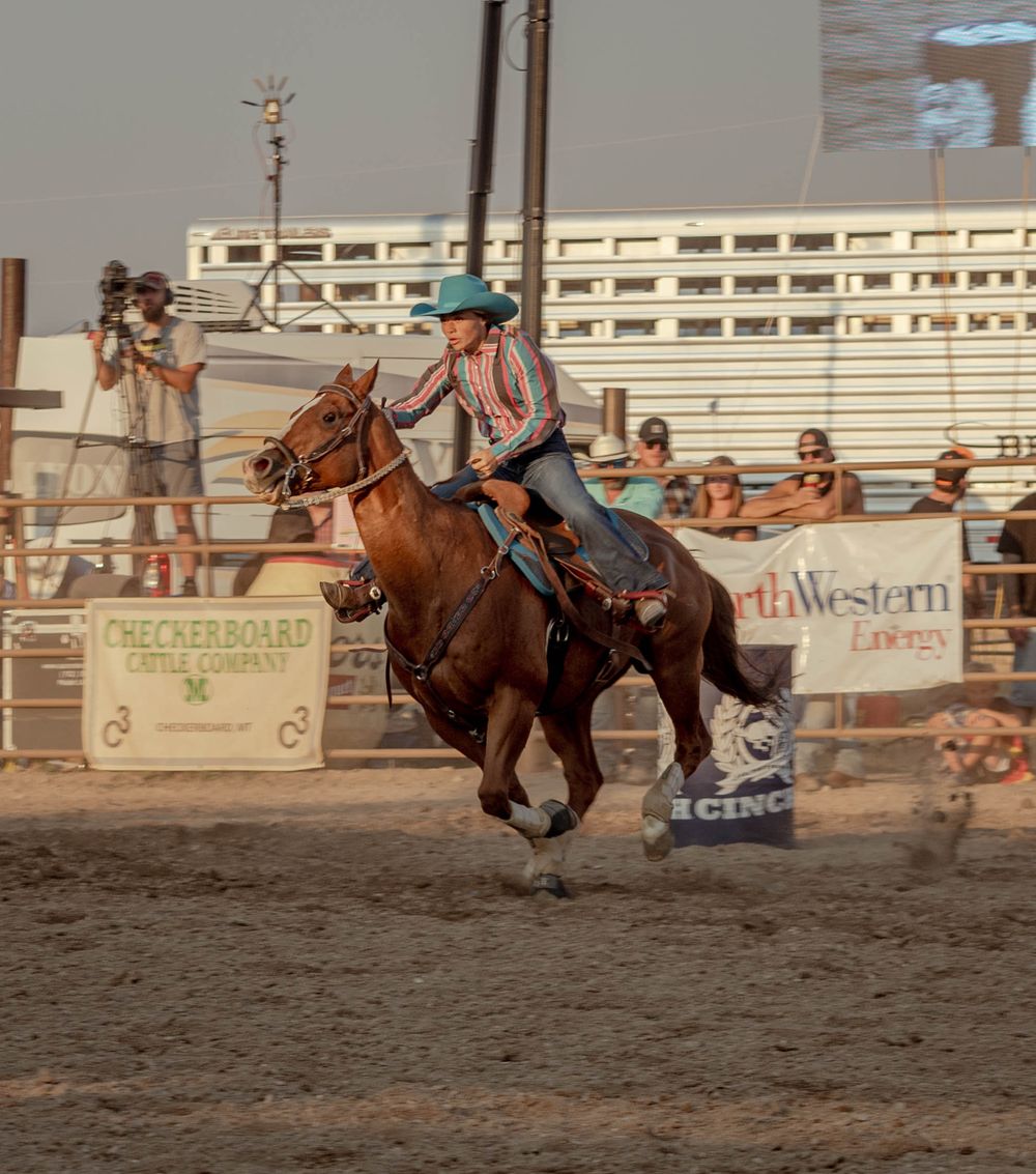 A cowgirl rides a galloping brown horse during a barrel racing competition at a rodeo arena.