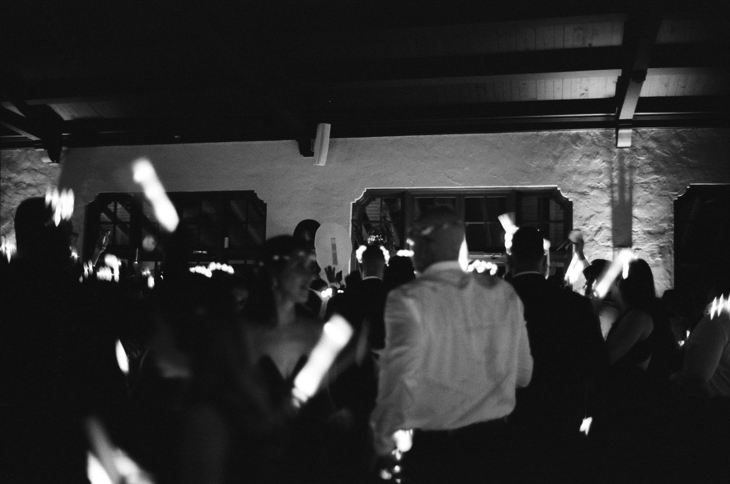 Black and white photo of a crowded indoor dance floor with guests holding glow sticks at a nighttime event.