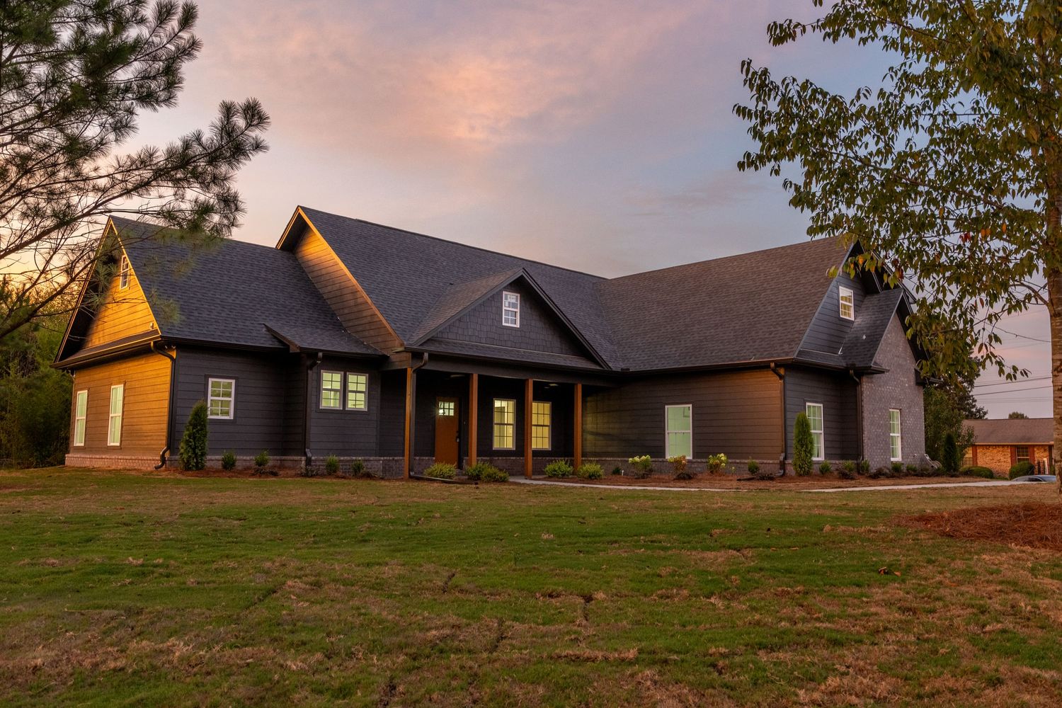 Craftsman-style ranch home at sunset with dark siding, covered porch, green lawn, and mature trees in a rural setting.
