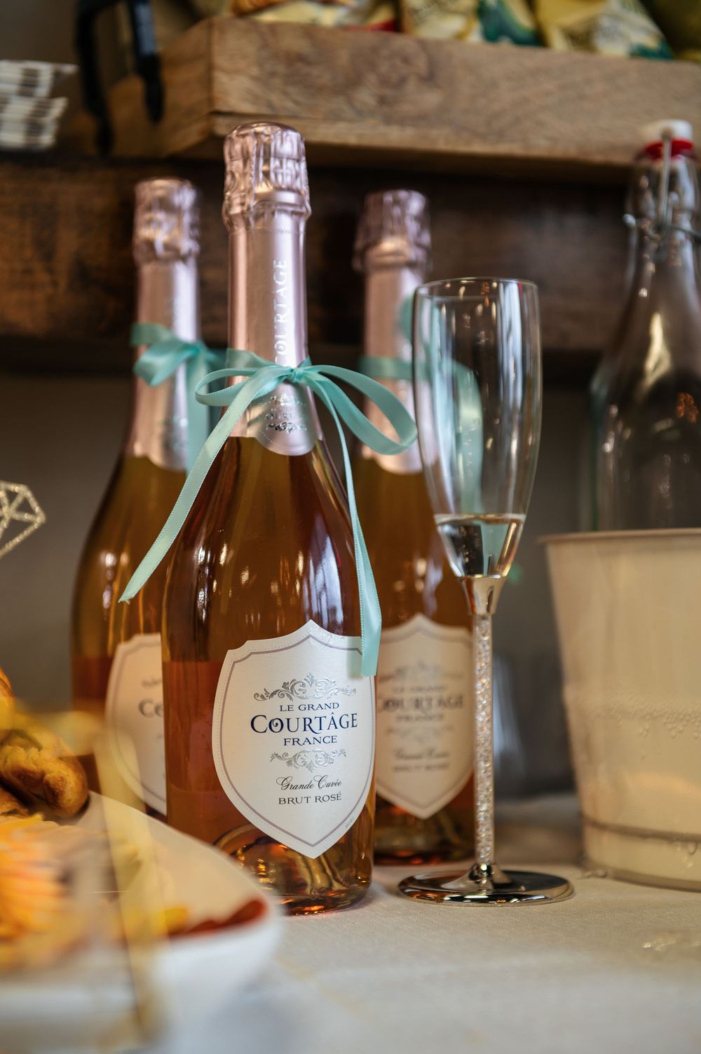 Bottles of Le Grand Courtage Brut Rose champagne with teal ribbon bow next to a crystal flute at an elegant celebration.