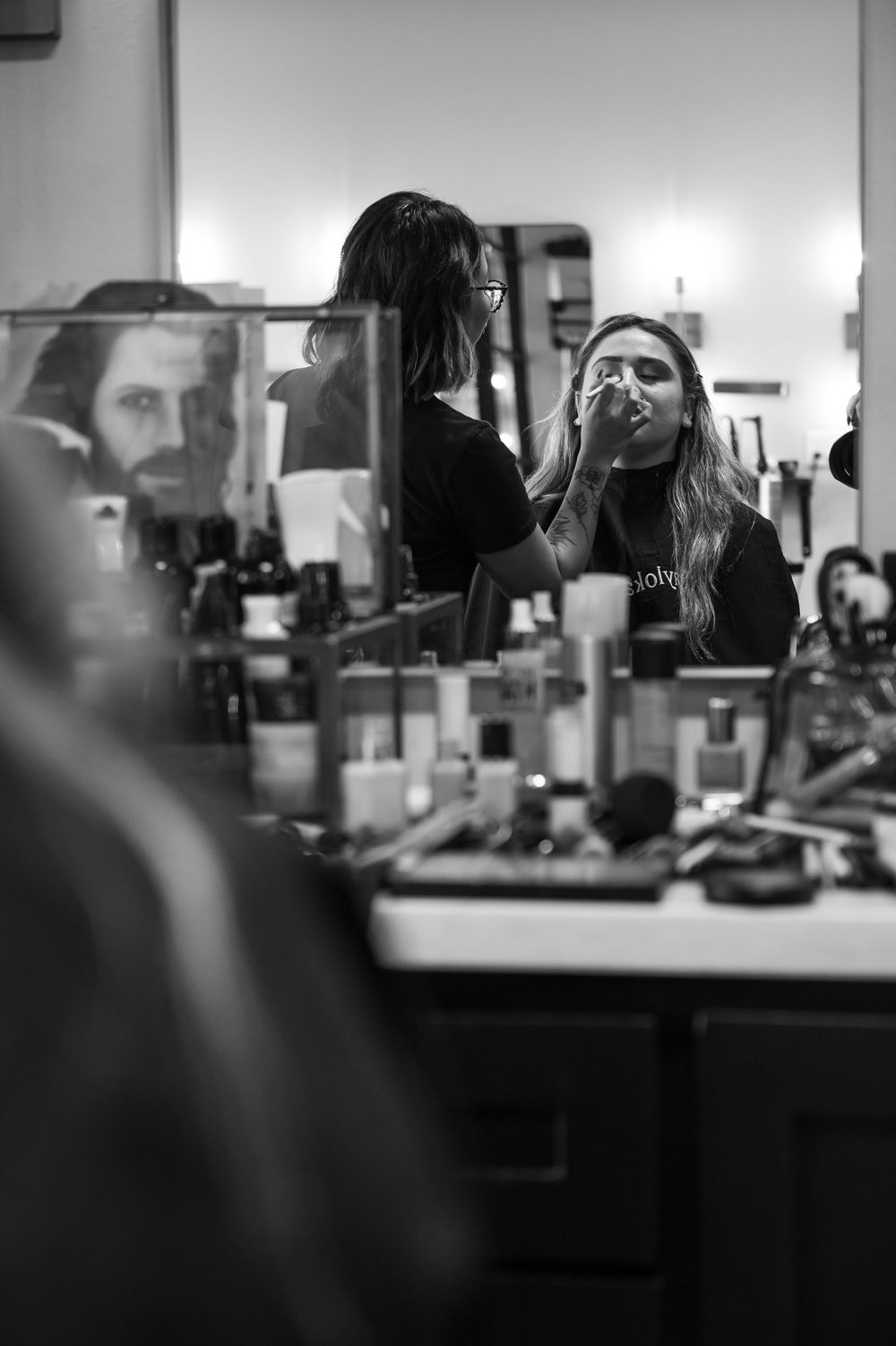 Black and white photo of a makeup artist applying cosmetics to a woman at a vanity mirror.