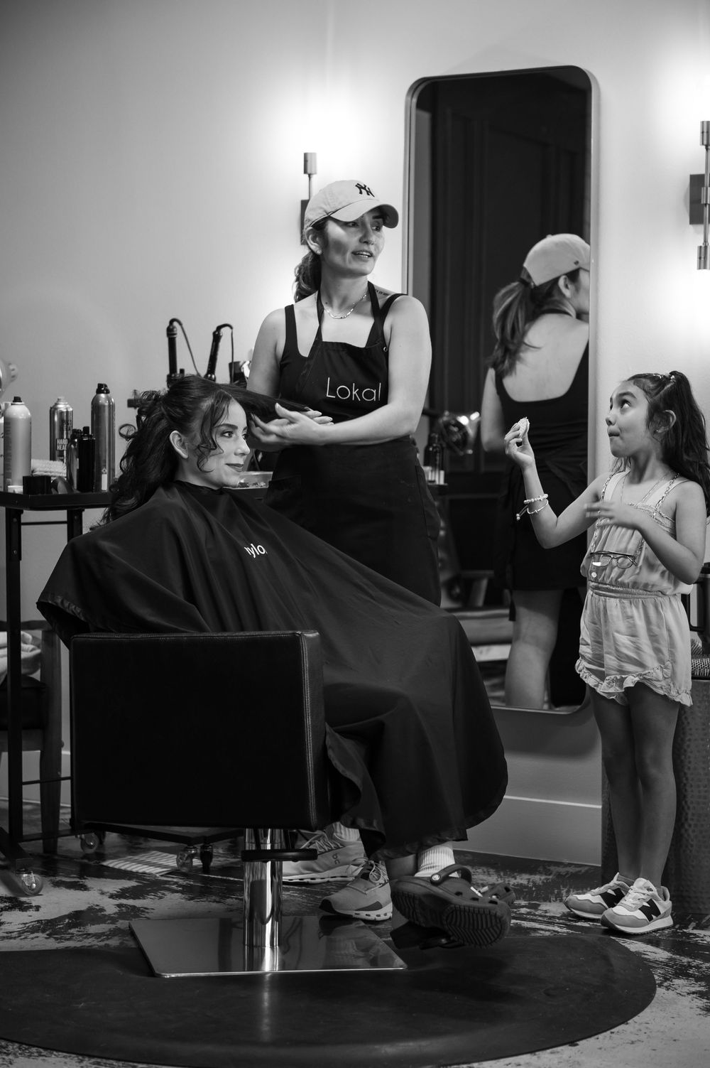 Hairstylist in Lokai apron smiling while styling a client in a salon chair, with a young girl standing nearby.