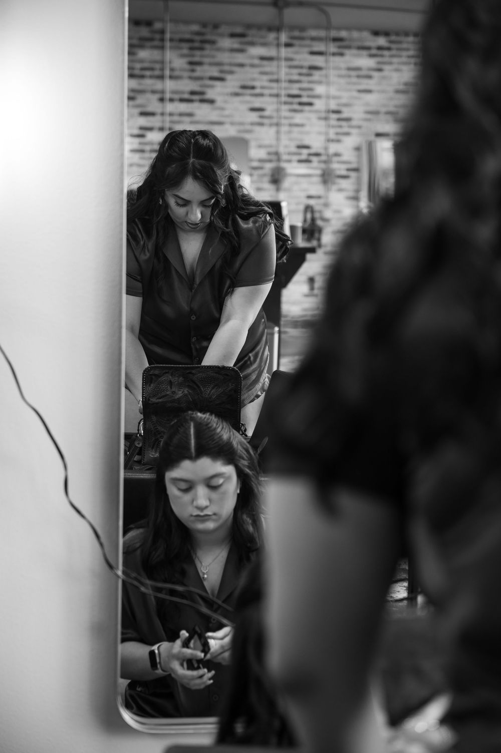 Black and white photo of a hairstylist working on a seated woman's hair in a salon mirror reflection.