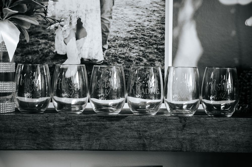 Six engraved stemless wine glasses displayed on a wooden shelf in black and white wedding photo.