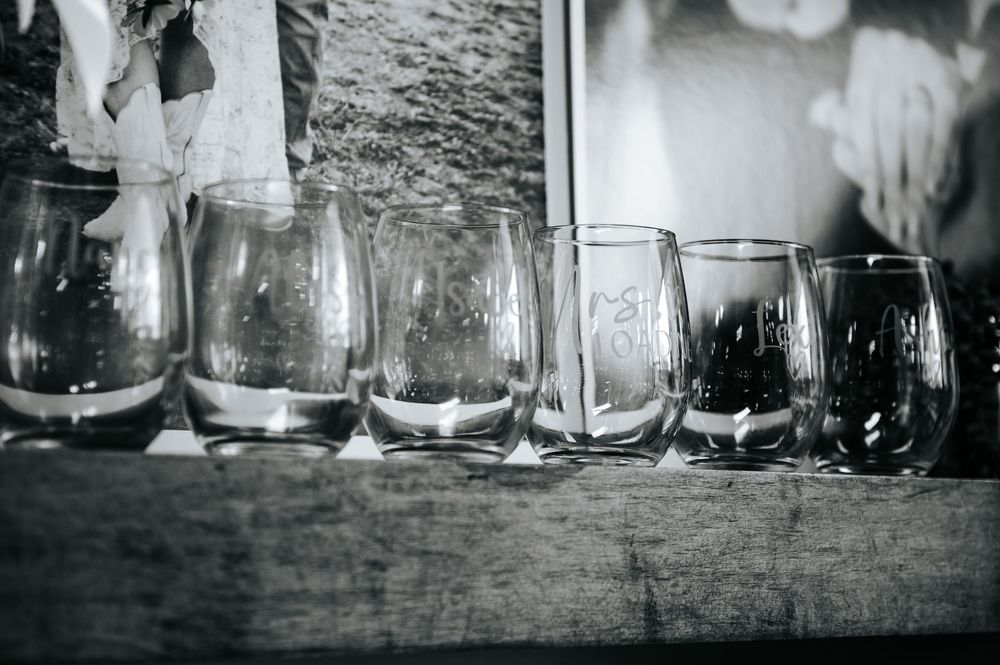 Black and white photo of various empty glass tumblers lined up on a rustic wooden shelf.