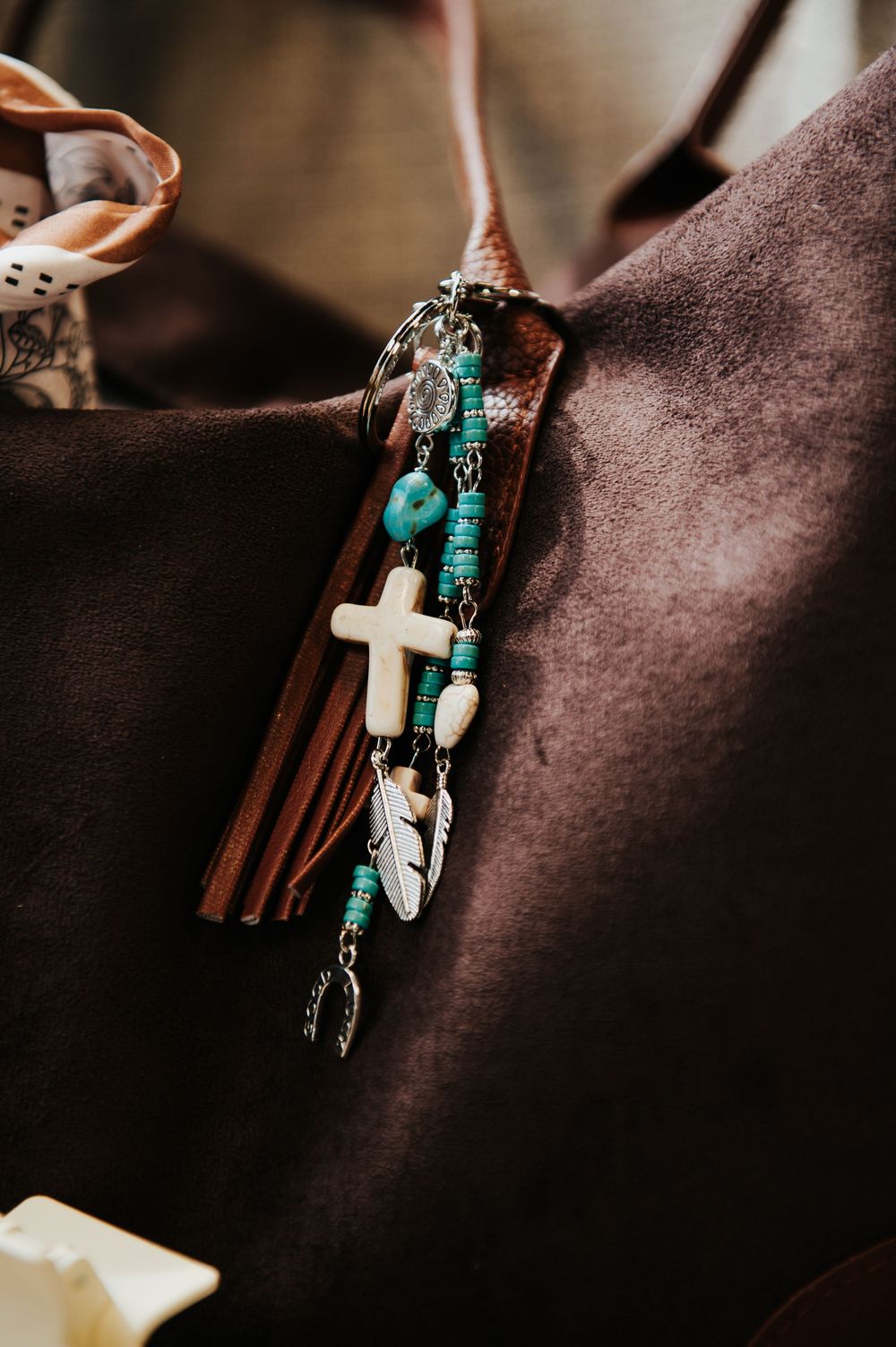 Boho western leather tassel keychain with white cross, turquoise beads, and feather charms on brown suede bag.