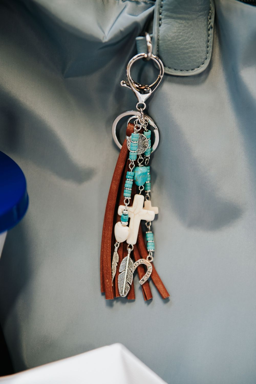 Boho keychain with turquoise beads, brown leather tassels, cross charm, and feather pendant on a gray handbag.