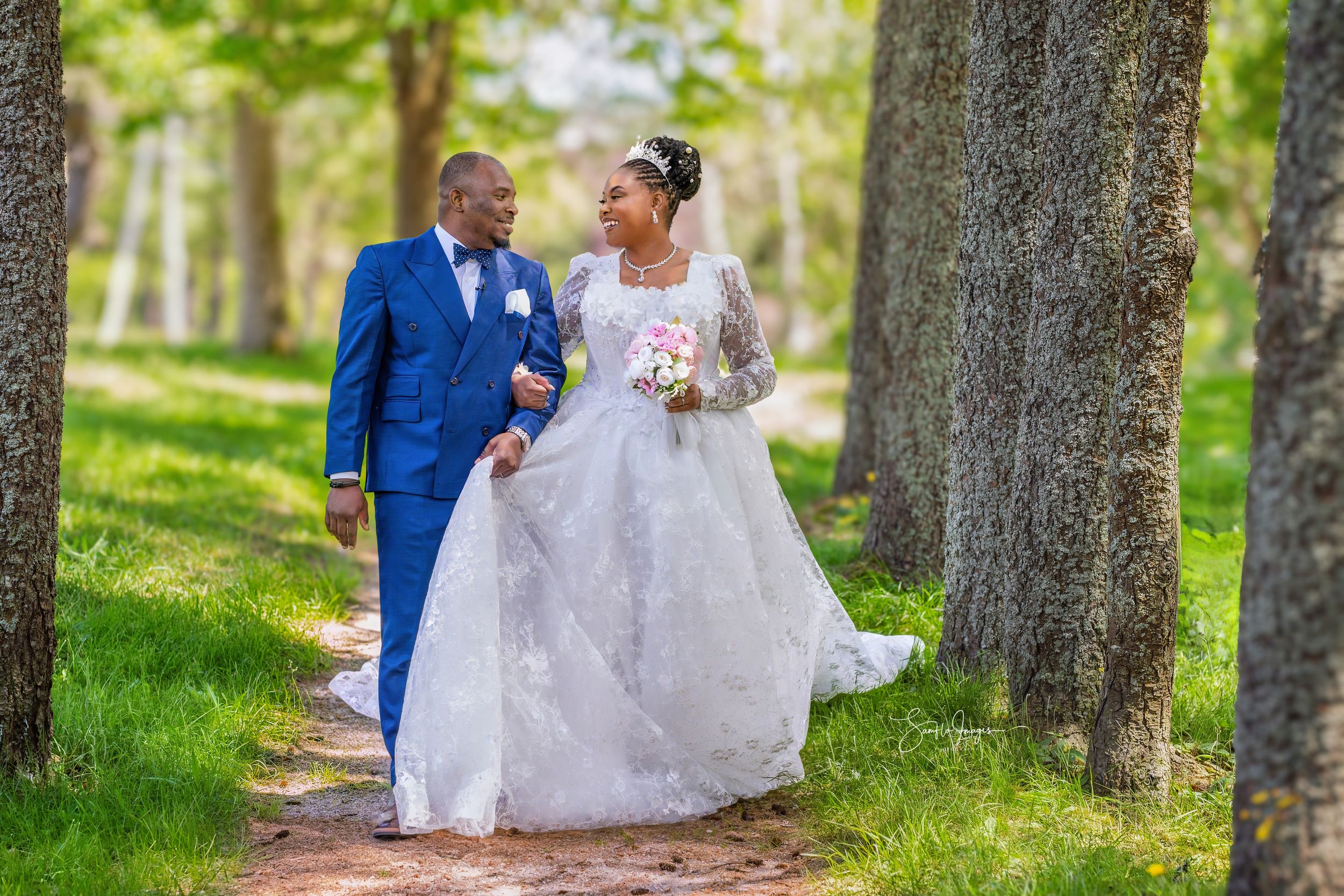 A couple shares a romantic kiss outdoors, groom in blue suit and bride in elegant white lace gown holding pink bouquet.