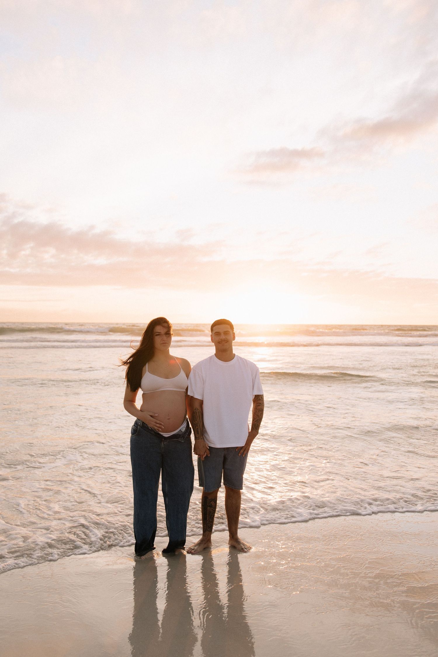 Pregnant woman in white maxi dress cradling baby bump on sandy beach at sunset.