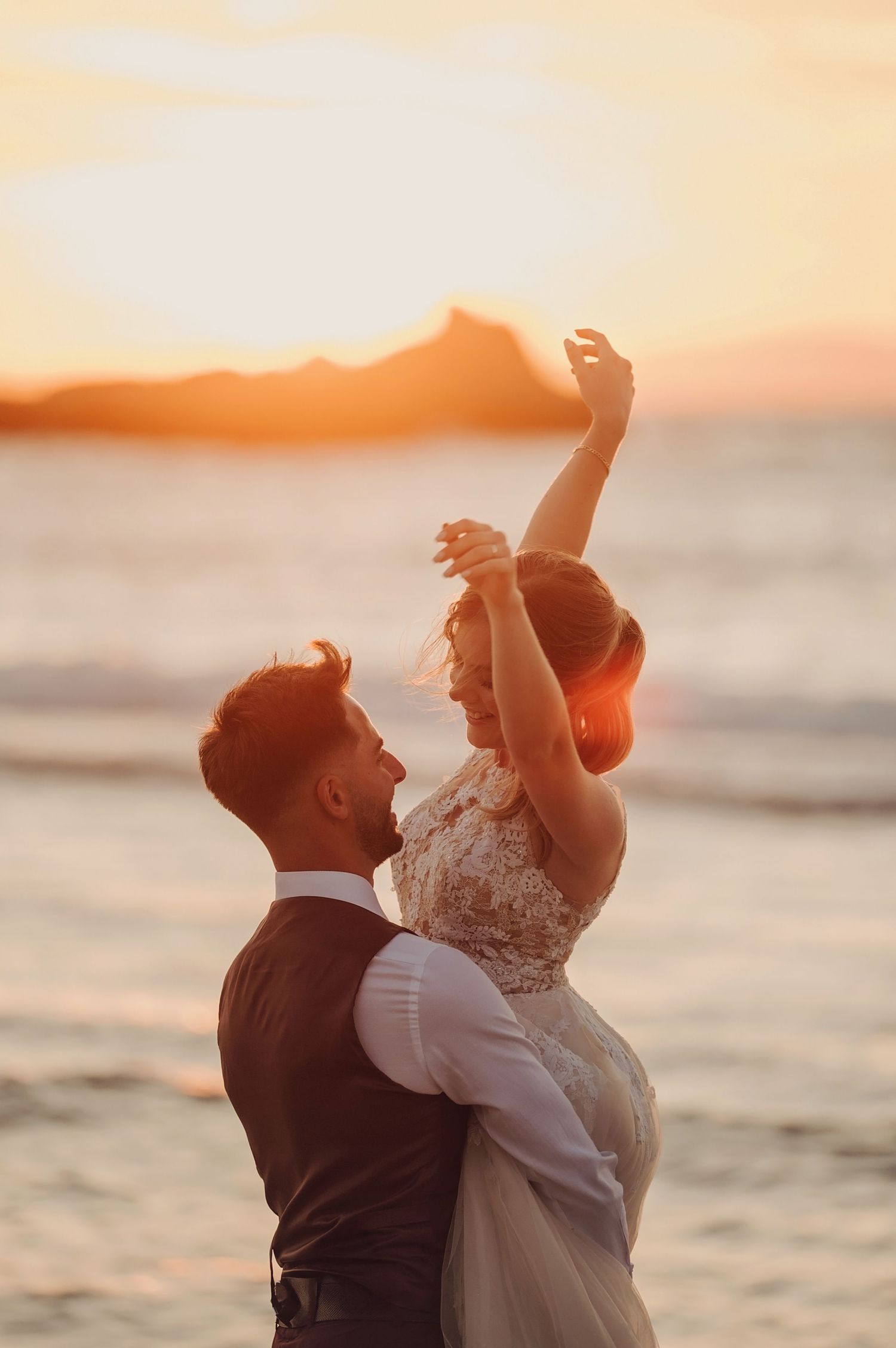 Couple dancing on beach at sunset, bride in lace dress with arm raised, groom in suit, golden hour ocean backdrop.
