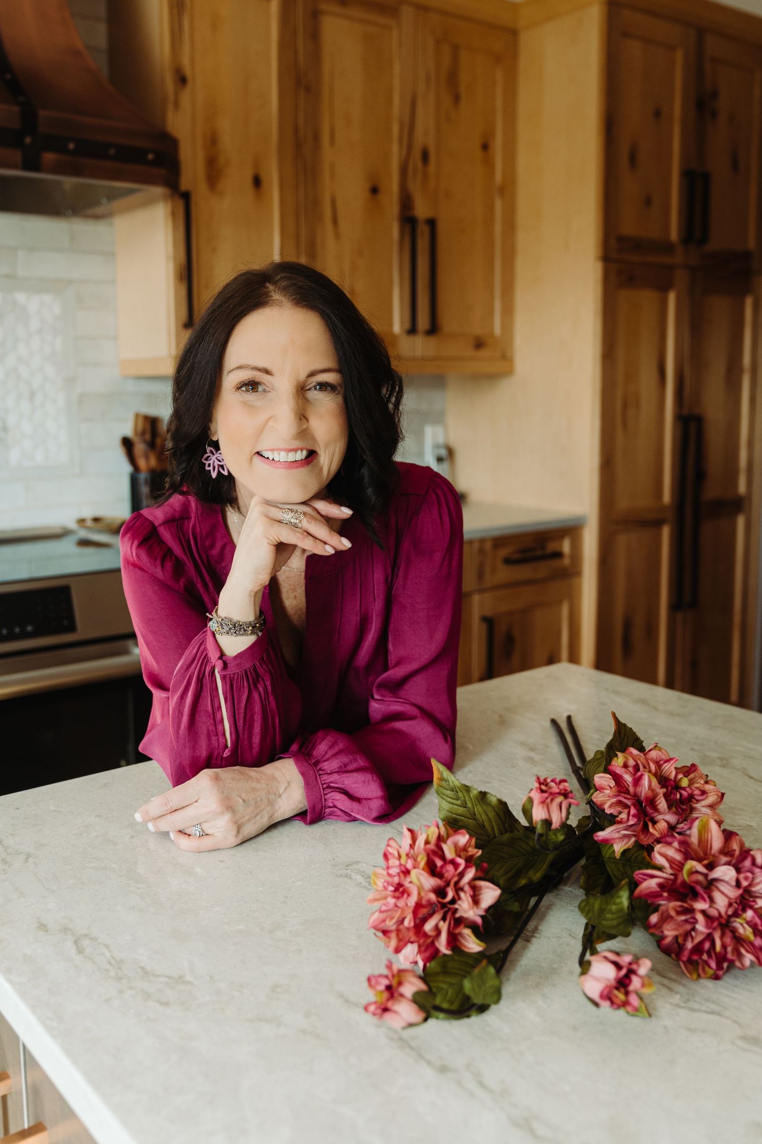 Smiling woman in magenta blouse leaning on marble kitchen counter with pink flowers in rustic wood kitchen.