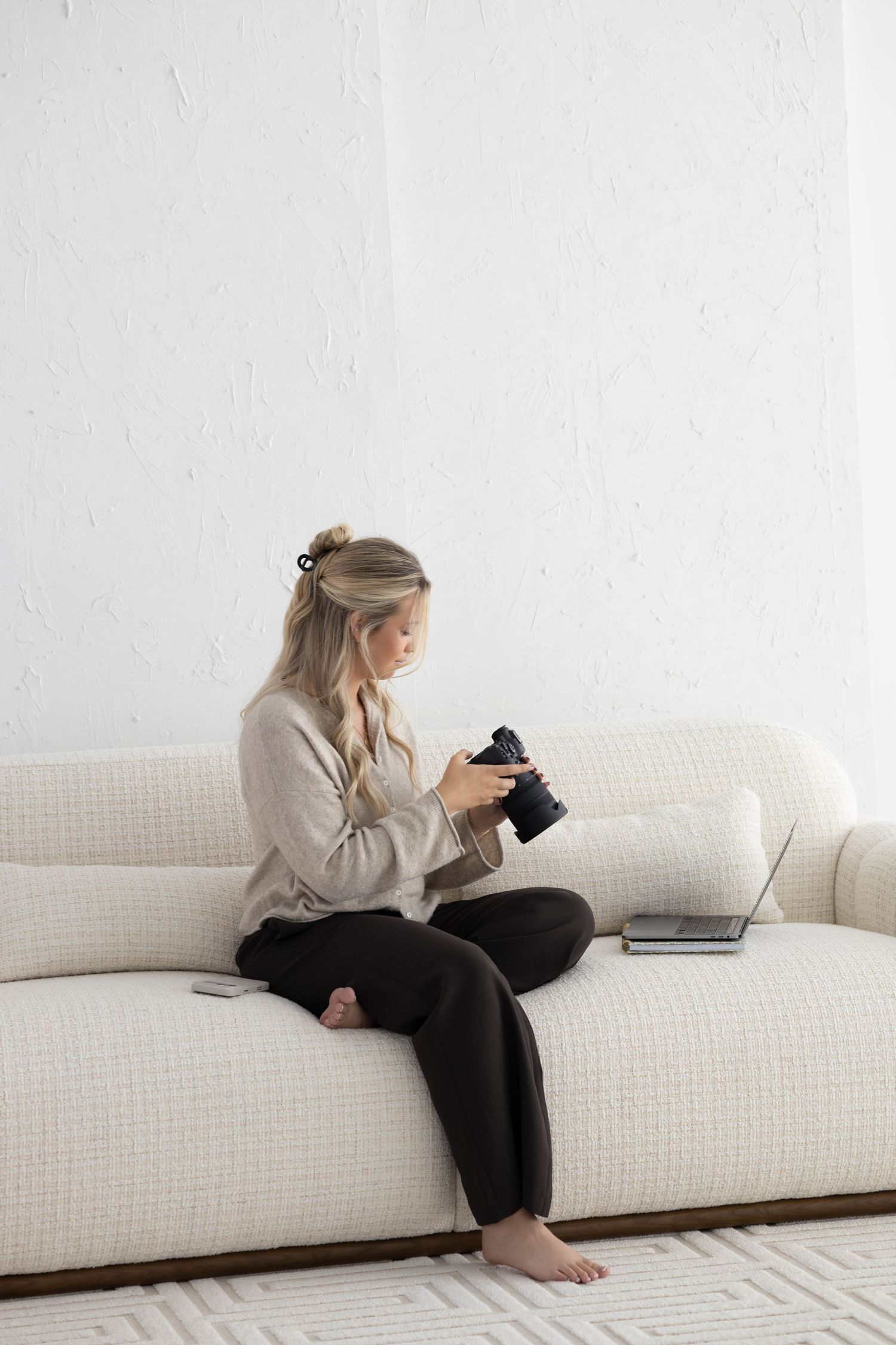 Woman smiling on white curved sofa with laptop and camera, beside arched wall and olive tree in bright modern studio.