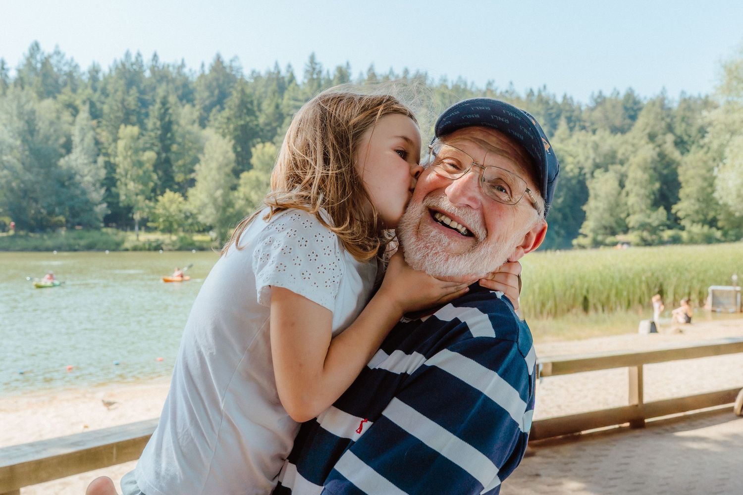 A young girl kisses an elderly man on the cheek near a lake surrounded by trees on a sunny day.