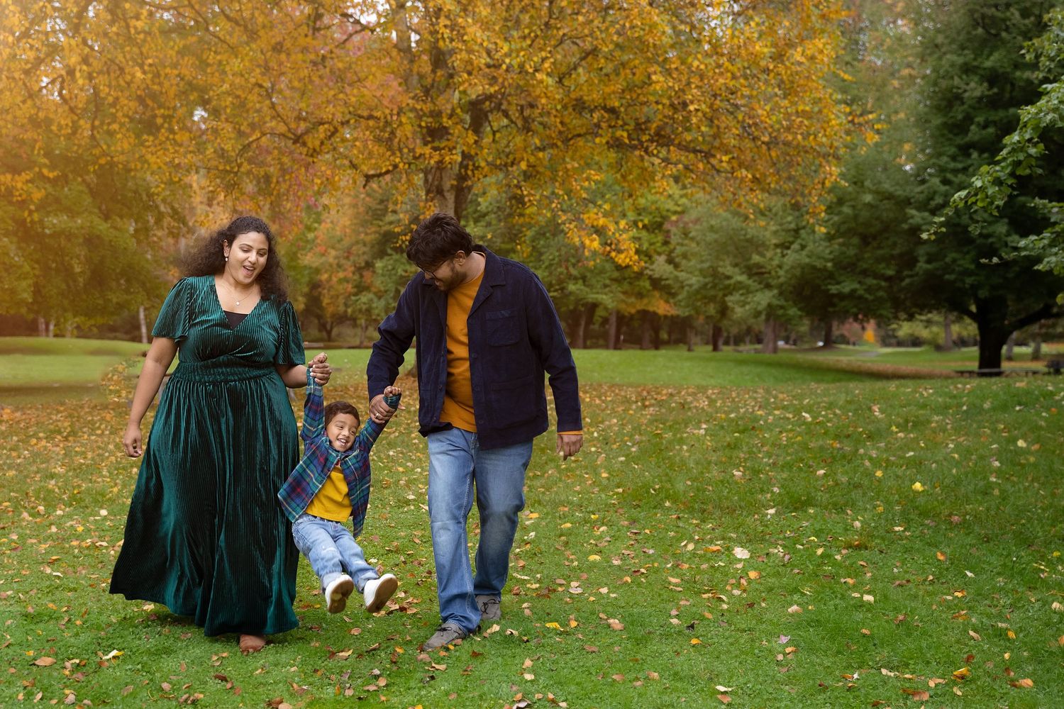 Family of three enjoying autumn walk in park, parents swinging young child between them on green grass.