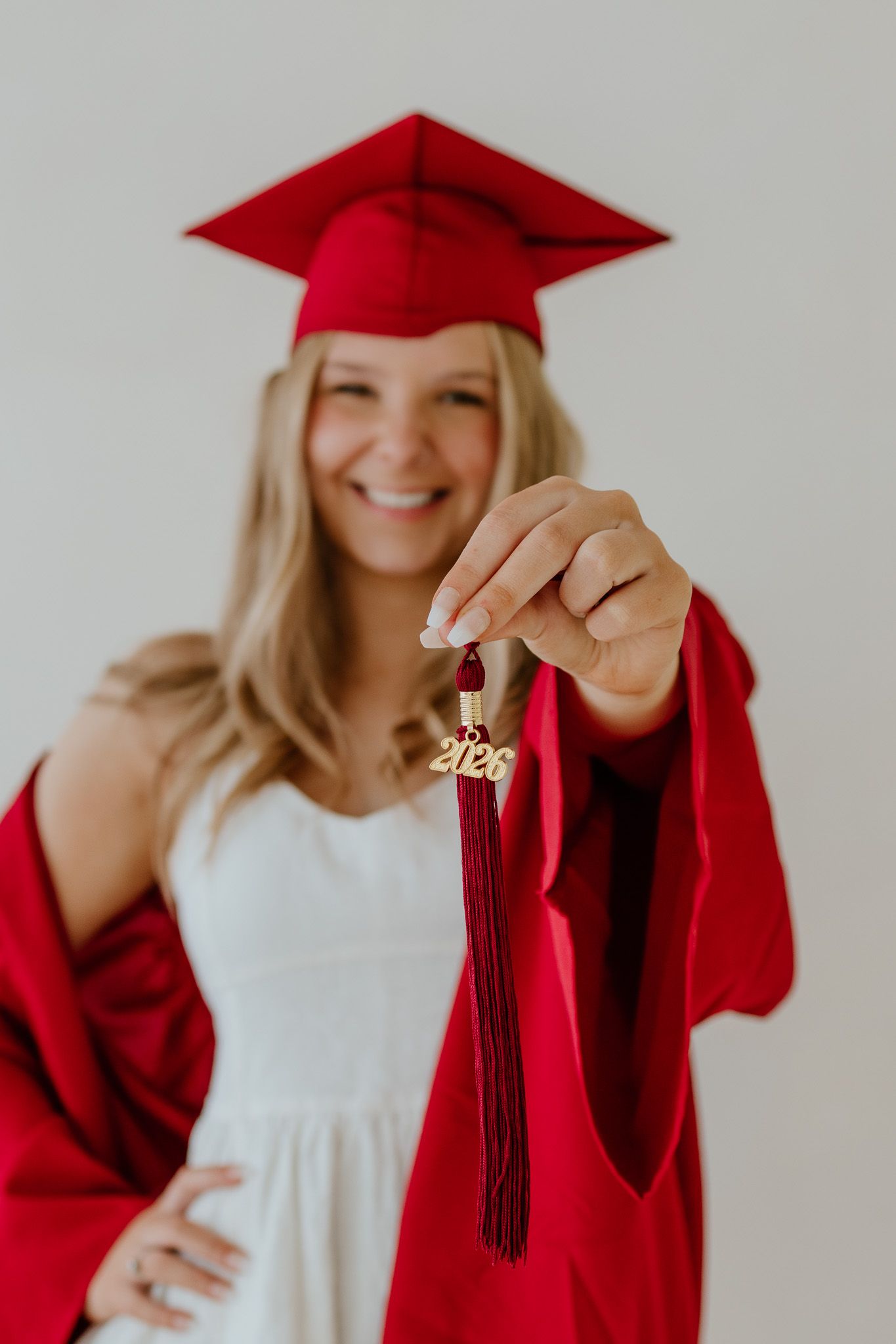 Smiling blonde graduate in red cap and gown holding a 2023 tassel close to camera during graduation photo session.